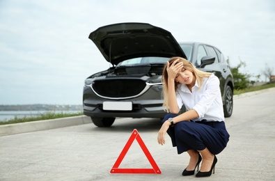 Photo of Upset woman sitting near warning triangle and broken car on road