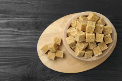 Photo of Brown sugar cubes in bowl on black wooden table, top view. Space for text