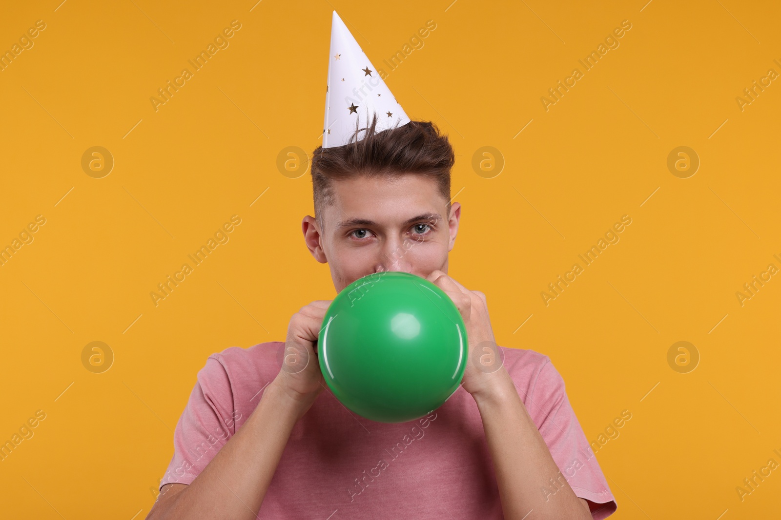 Photo of Young man in party hat inflating balloon on orange background