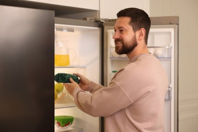 Photo of Man putting bowl covered with beeswax food wrap into refrigerator indoors