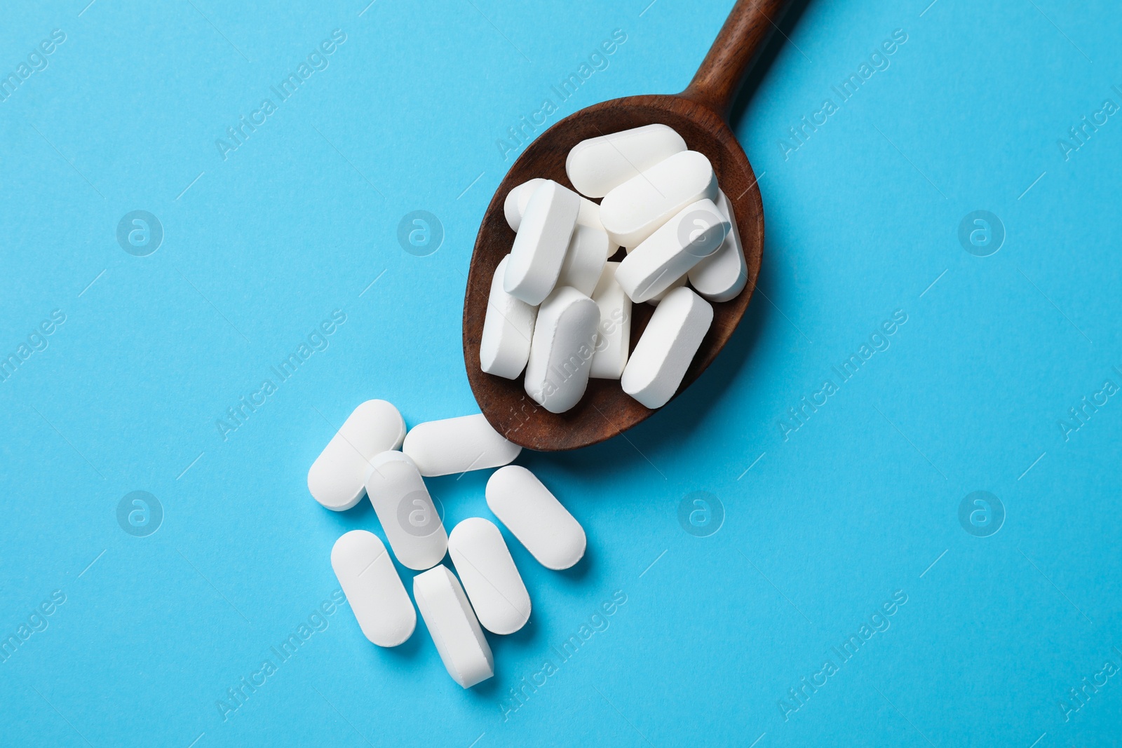 Photo of Vitamin capsules in wooden spoon on light blue background, top view