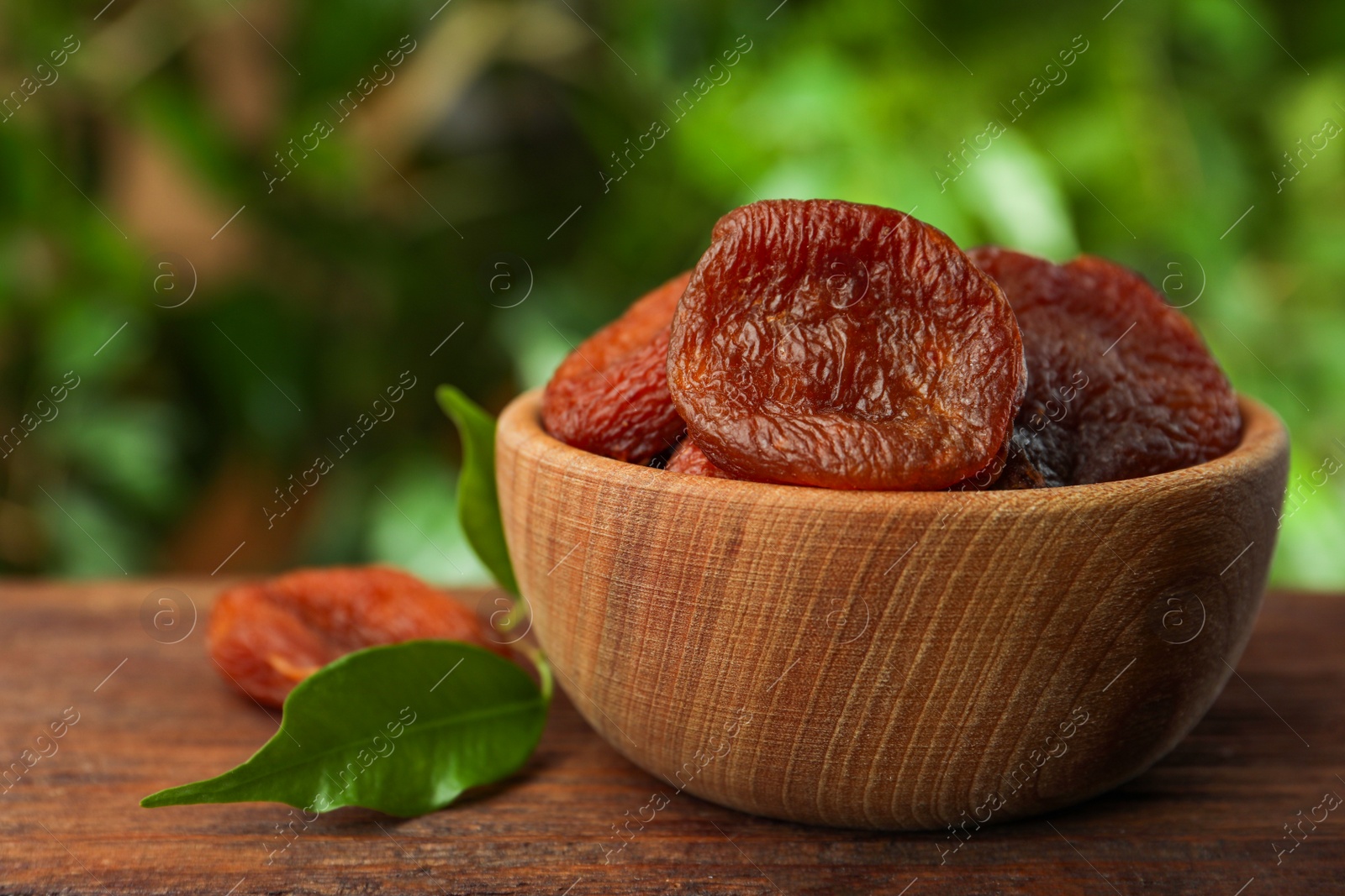 Photo of Bowl of tasty apricots on wooden table against blurred green background. Dried fruits