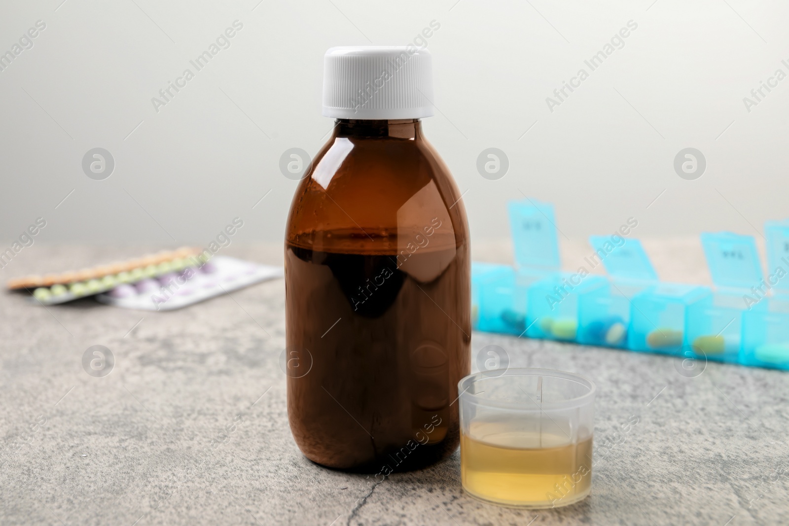 Photo of Bottle of syrup and measuring cup on light grey table. Cold medicine