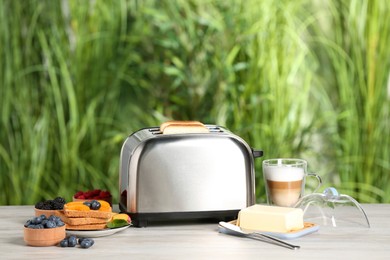 Photo of Modern toaster and fresh products on white wooden table against blurred green background