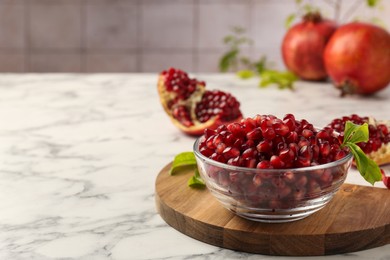 Photo of Ripe juicy pomegranate grains in bowl and green leaves on white marble table, space for text