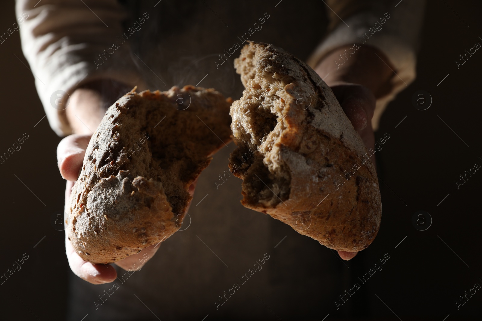 Photo of Man breaking loaf of fresh bread on dark background, closeup