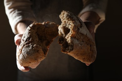 Photo of Man breaking loaf of fresh bread on dark background, closeup