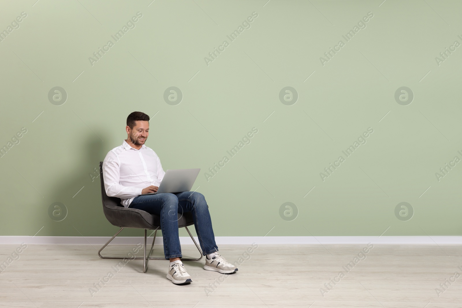 Photo of Happy man with laptop sitting in armchair near light green wall indoors, space for text