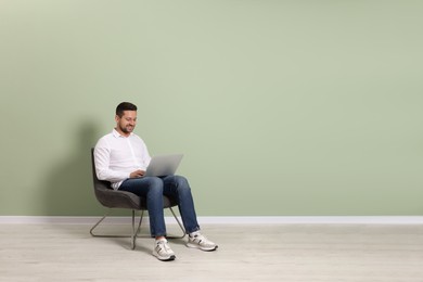 Happy man with laptop sitting in armchair near light green wall indoors, space for text