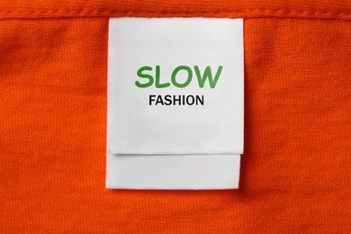 Image of Conscious consumption. Clothing label with words Slow Fashion on orange garment, top view
