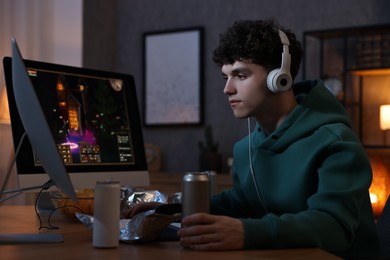 Photo of Young man with energy drink and headphones playing video game at wooden desk indoors