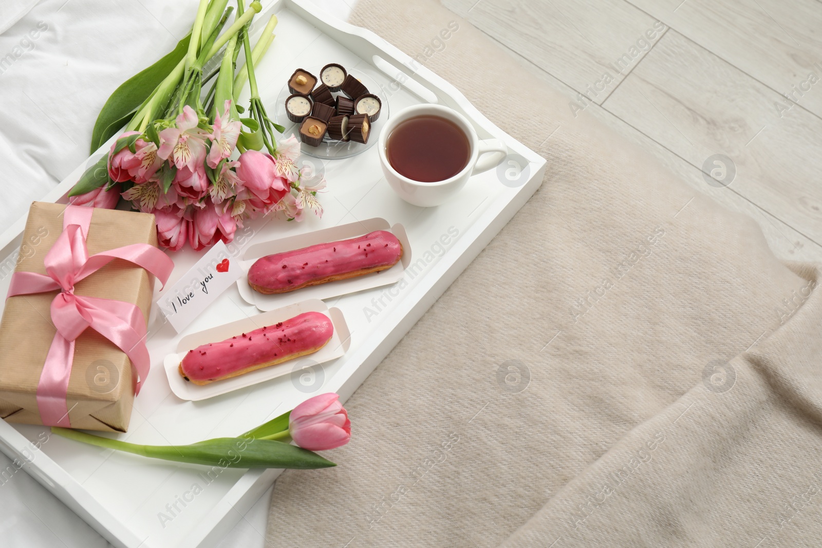 Photo of Tasty breakfast served in bed. Delicious desserts, tea, flowers, gift box and card with phrase I Love You on tray, above view. Space for text