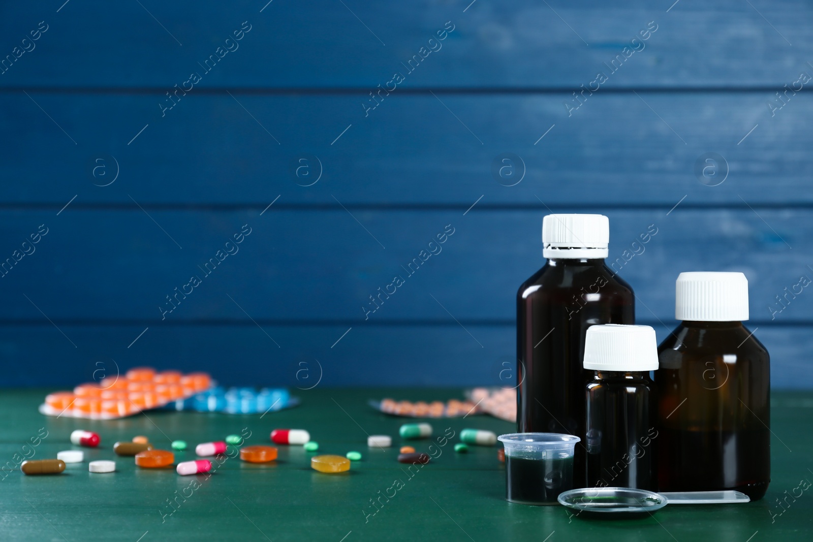 Photo of Bottles of cough syrup, dosing spoon, measuring cup and pills on green wooden table. Space for text