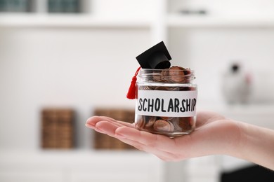 Photo of Woman holding glass jar of coins and graduation cap indoors, closeup with space for text. Scholarship concept