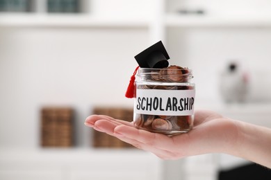 Woman holding glass jar of coins and graduation cap indoors, closeup with space for text. Scholarship concept