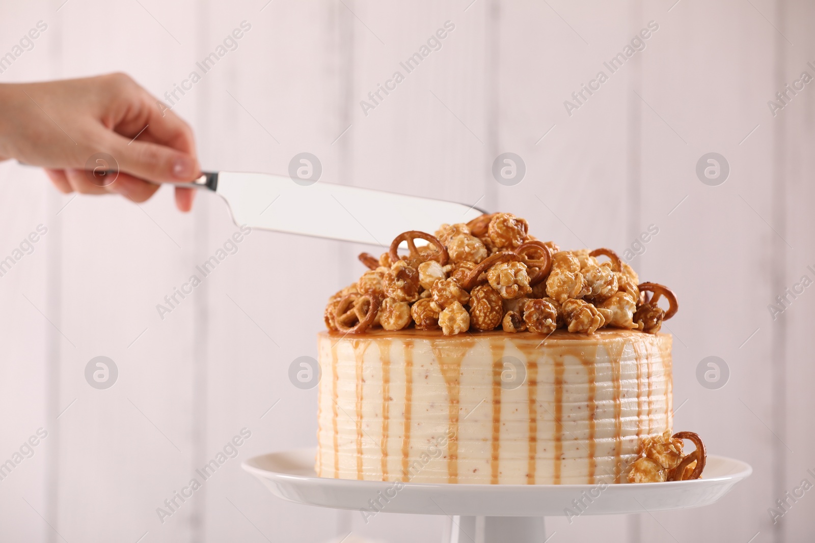 Photo of Woman cutting caramel drip cake decorated with popcorn and pretzels against light background, closeup