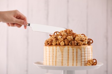 Woman cutting caramel drip cake decorated with popcorn and pretzels against light background, closeup