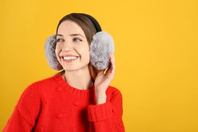 Happy woman wearing warm earmuffs on yellow background, space for text