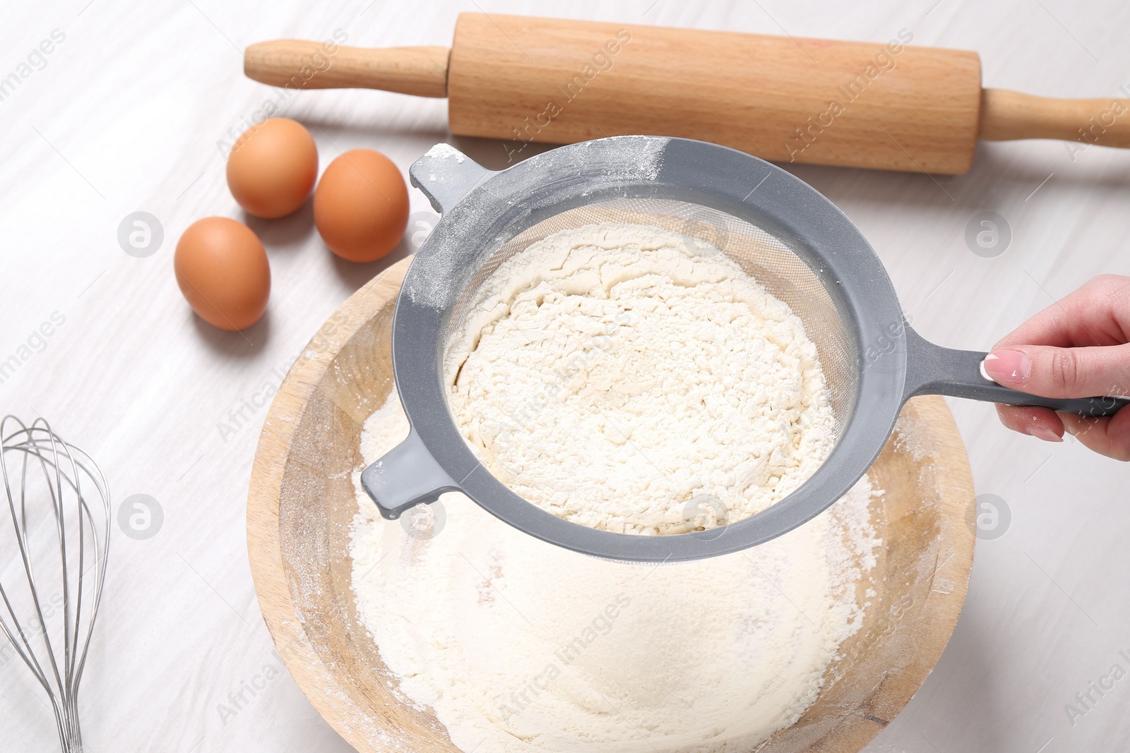 Photo of Woman sieving flour into bowl at white wooden table, above view