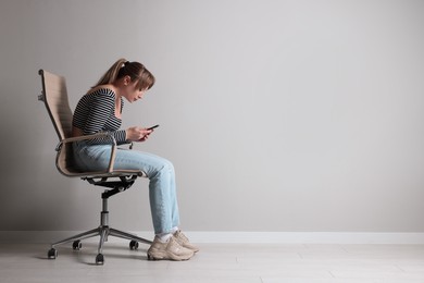 Photo of Young woman with poor posture using smartphone while sitting on chair near grey wall, space for text