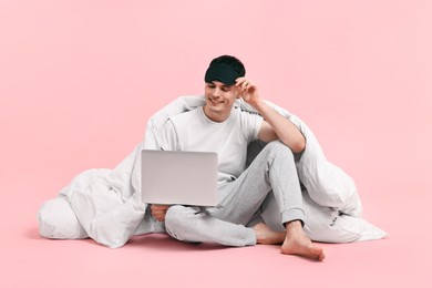 Photo of Happy man in pyjama with sleep mask, blanket and laptop on pink background