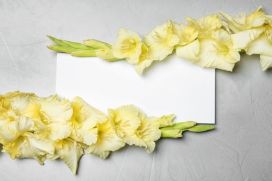 Photo of Beautiful gladiolus flowers with blank card on gray background, top view