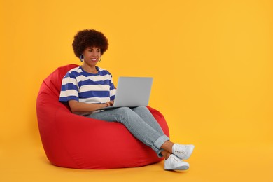 Beautiful young woman with laptop sitting on beanbag chair against orange background. Space for text