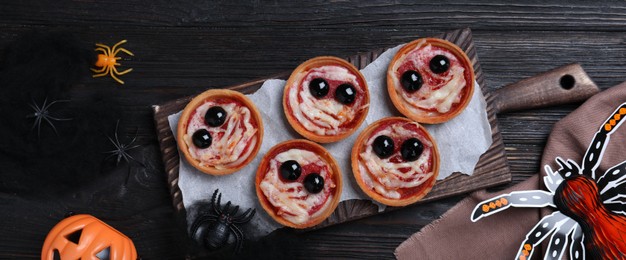 Photo of Cute monster tartlets served on black wooden table, flat lay with space for text. Halloween party food