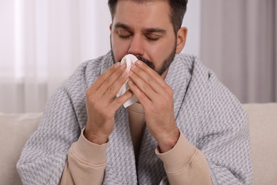 Photo of Sick man wrapped in blanket with tissue blowing nose on sofa indoors. Cold symptoms