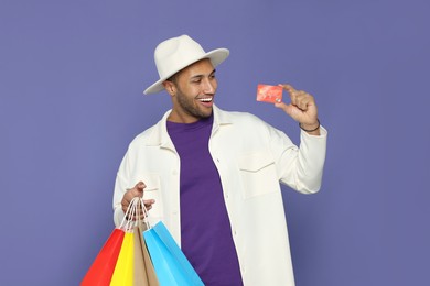 Happy African American man in hat with shopping bags and credit card on purple background
