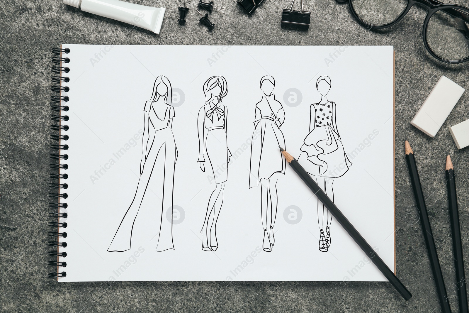 Image of Sketches of different clothes in pad on grey textured table. Fashion designer's desk with stationery, flat lay