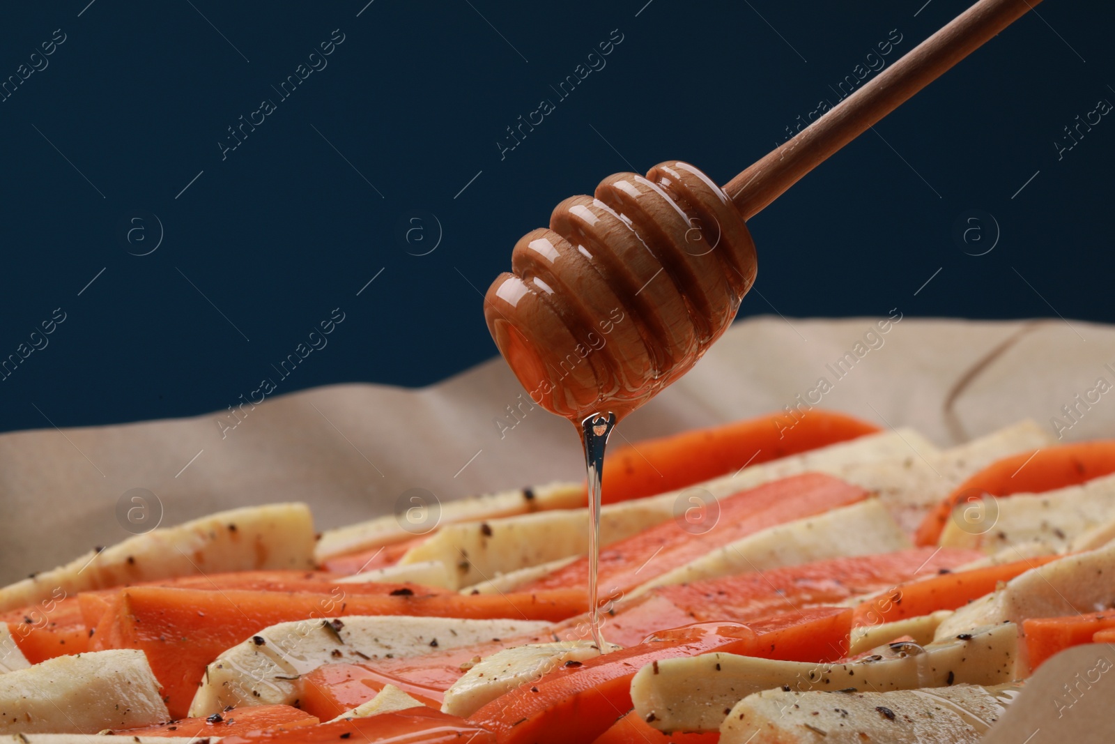 Photo of Pouring honey onto slices of parsnip and carrot against blue background, closeup