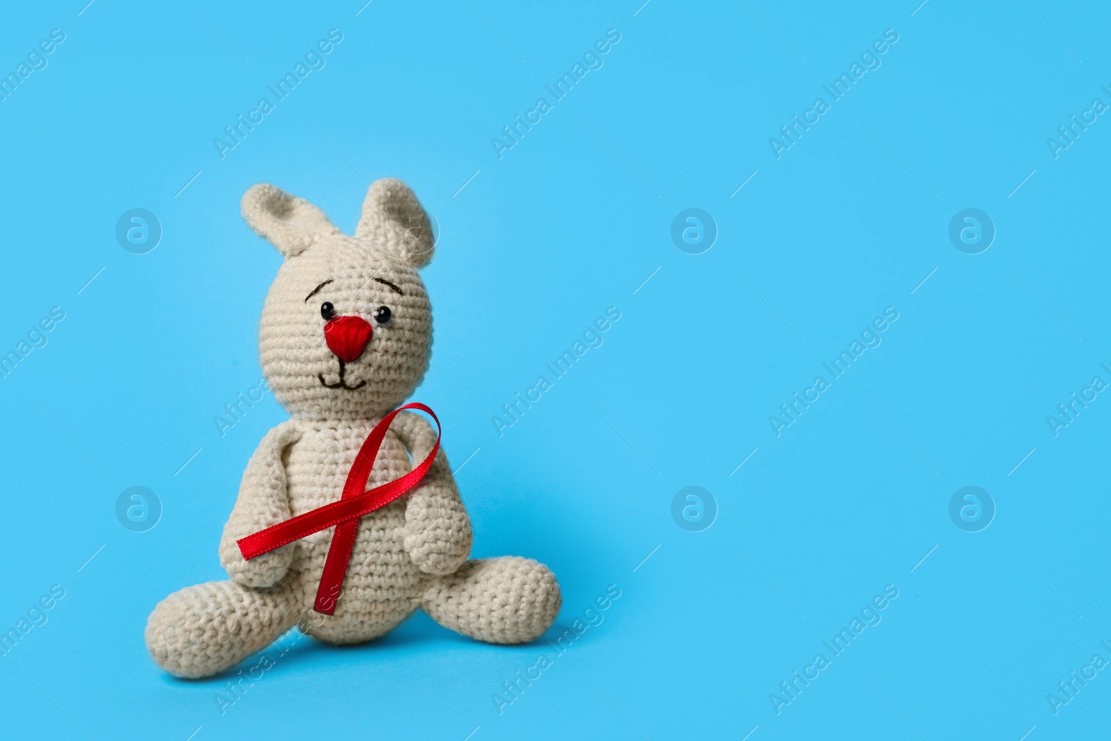 Photo of Cute knitted toy bunny with red ribbon on blue background, space for text. AIDS disease awareness