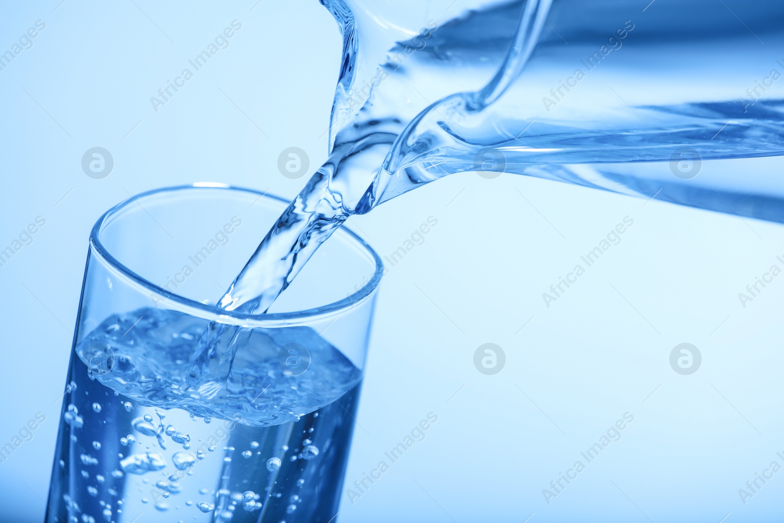 Photo of Pouring water from jug into glass on light blue background, closeup