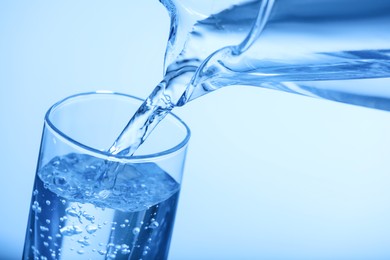Pouring water from jug into glass on light blue background, closeup