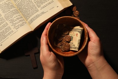 Donate and give concept. Woman with bowl of money, closeup. Bible and cross on black wooden table, top view