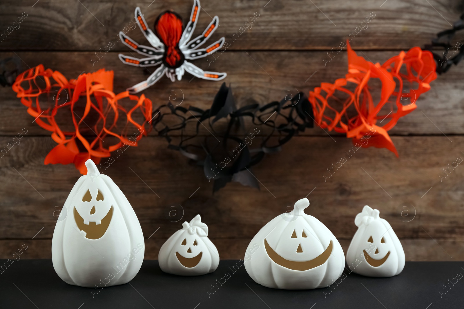 Image of White pumpkin shaped candle holders on black table against wooden wall with paper spider and garland. Halloween decoration