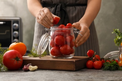 Photo of Woman putting tomatoes into glass jar at grey kitchen table, closeup. Pickling vegetables