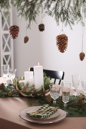 Photo of Christmas celebration. Cones hanging from fir tree branch over table with burning candles and tableware