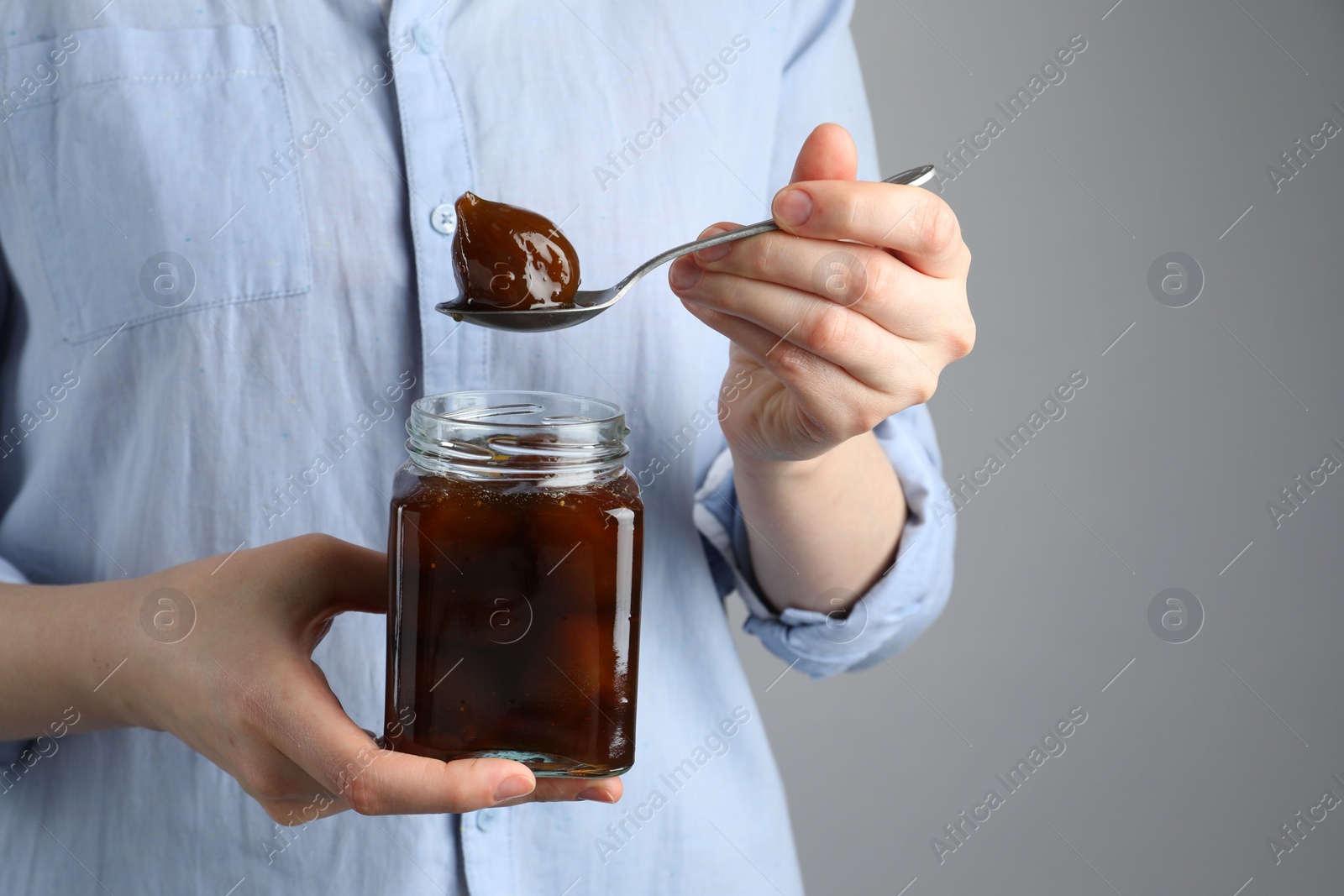 Photo of Woman holding jar of tasty sweet fig jam on grey background, closeup. Space for text
