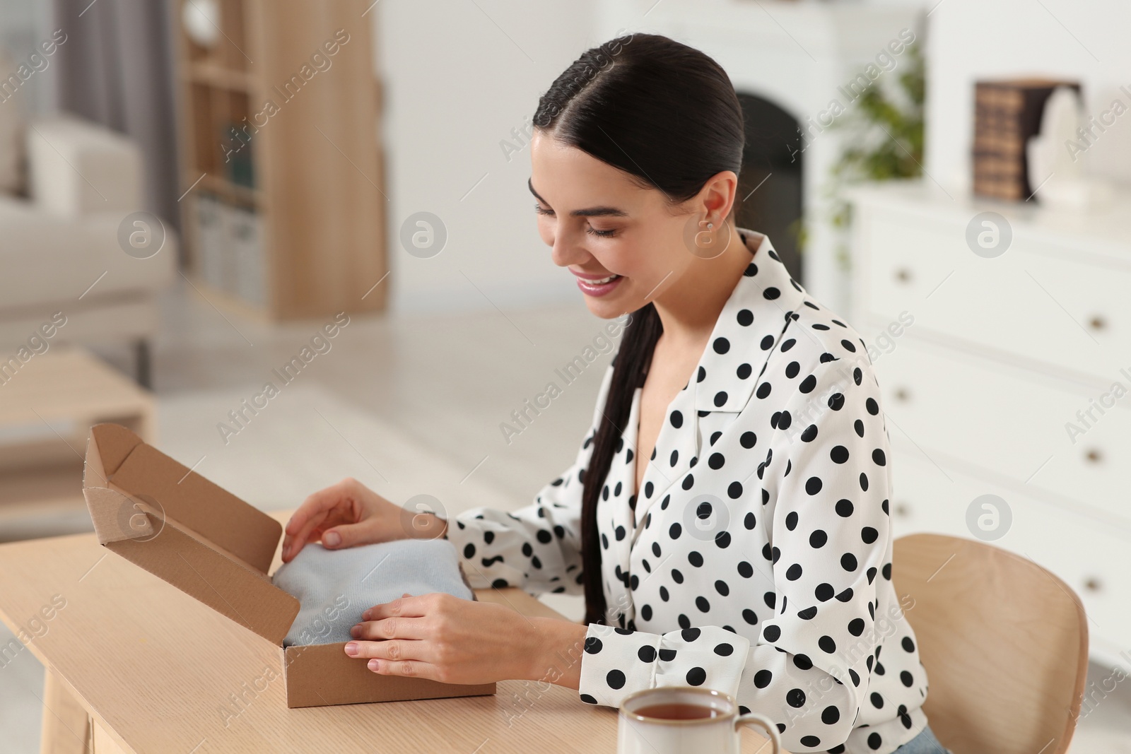 Photo of Happy young woman opening parcel at table indoors. Internet shopping