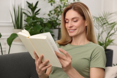 Photo of Woman reading book on sofa surrounded by beautiful potted houseplants at home