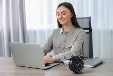 Photo of Young woman working with laptop at table, focus on alarm clock
