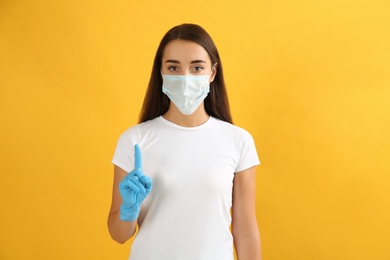 Woman in protective face mask and medical gloves with raised index finger on yellow background