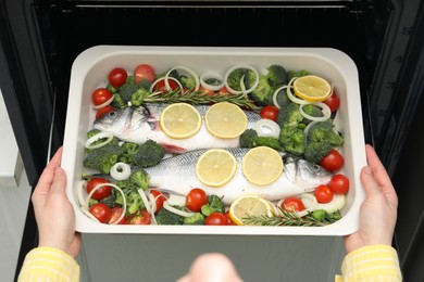 Photo of Woman putting baking dish with raw fish and vegetables into oven, top view