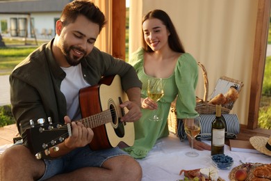 Romantic date. Beautiful woman with glass of wine and her boyfriend playing guitar during picnic in wooden gazebo