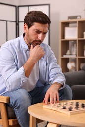 Thoughtful man playing checkers in armchair at home