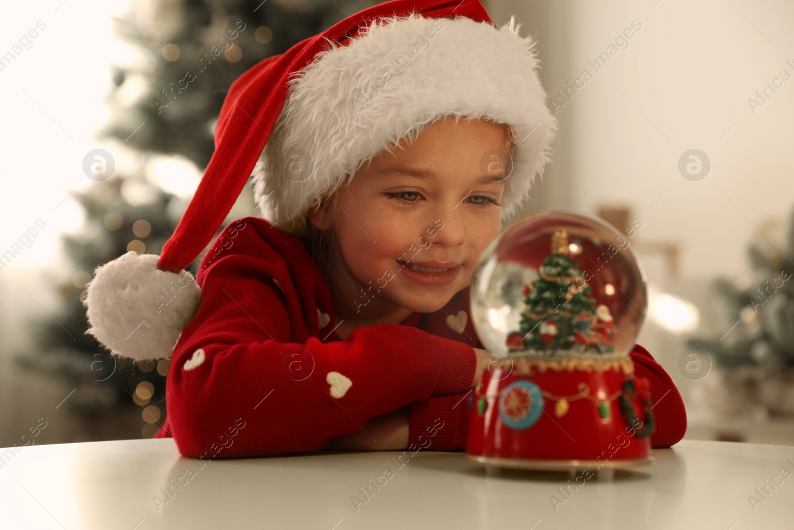 Photo of Little girl in Santa hat playing with snow globe at home