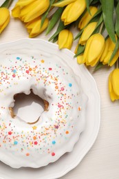 Photo of Easter cake with sprinkles and tulips on white wooden table, flat lay