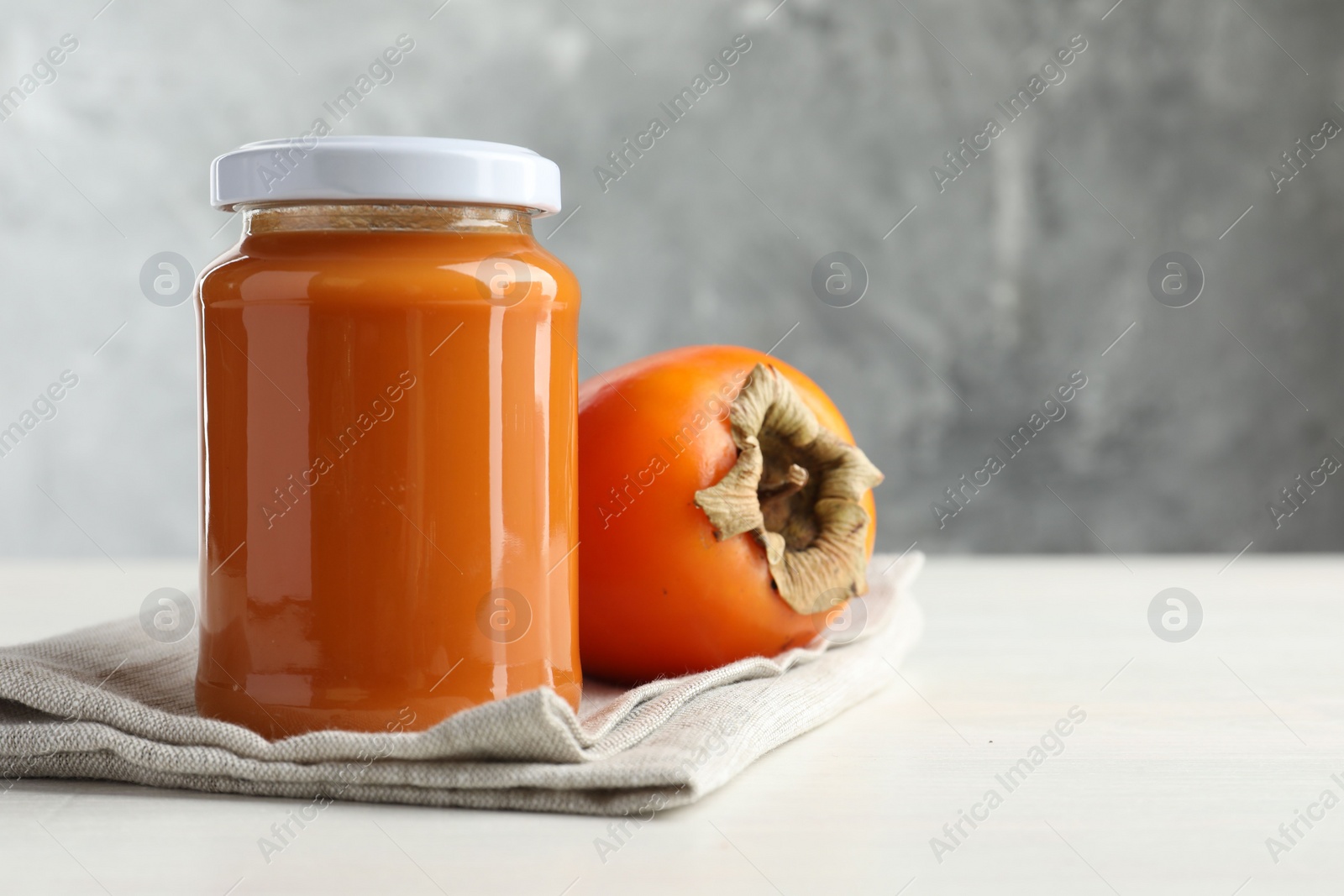 Photo of Delicious persimmon jam in glass jar and fresh fruit on white wooden table, space for text